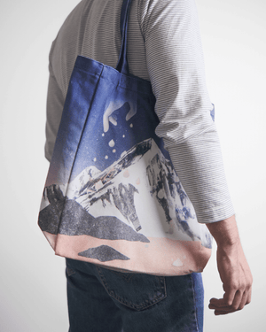 A person wearing a gray striped long-sleeve shirt and blue jeans carries a canvas tote bag over their shoulder. The tote bag features a scenic print of snow-capped mountains under a starry night sky with a soft pink horizon.