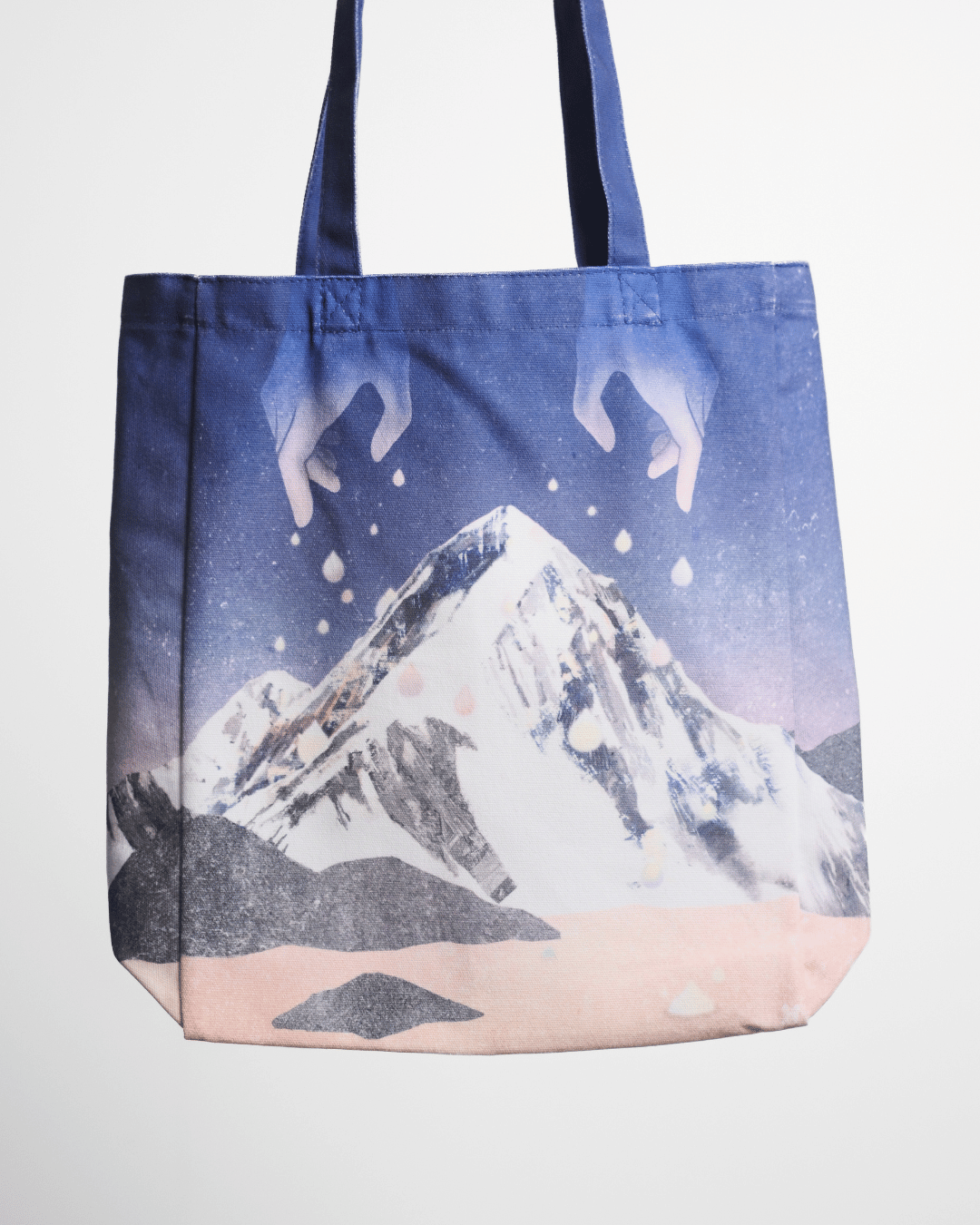 A canvas tote bag featuring a printed graphic of a snow-covered mountain under a starry blue and purple sky, with pale hands reaching down from above and a soft pink base. The bag has dark blue handles and is photographed against a plain white background.