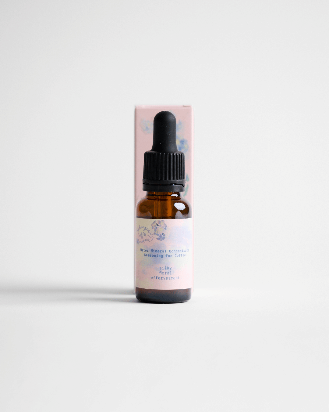 A small amber glass dropper bottle standing in front of a pale pink rectangular box on a clean white studio background.