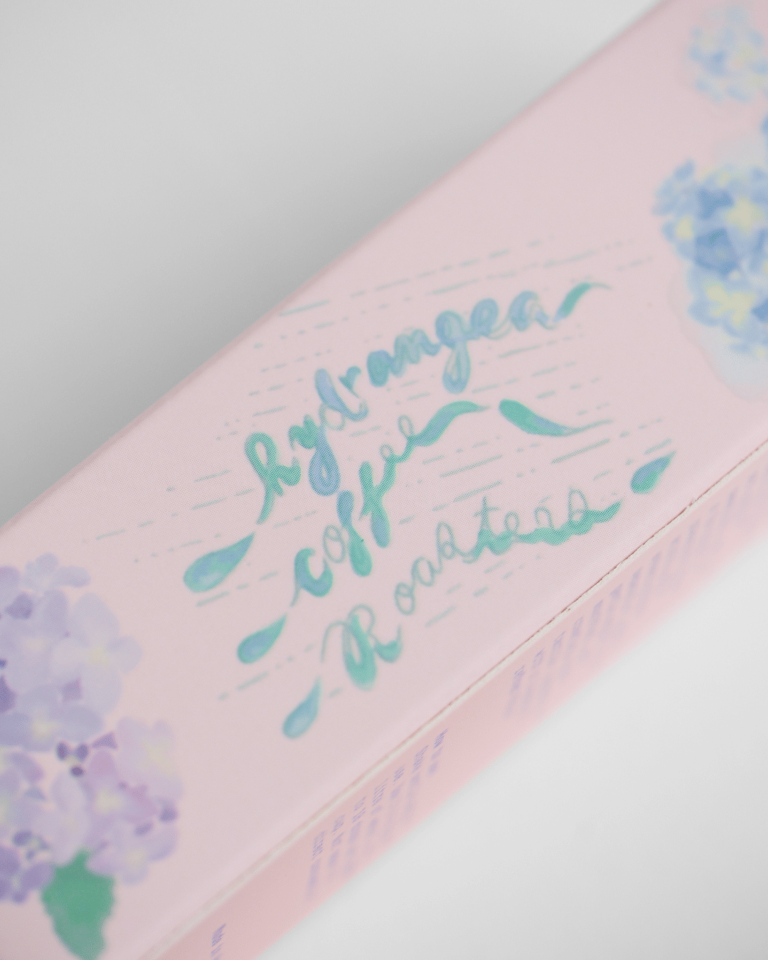 A close-up of a pink product box with teal cursive text reading 'Hydrangea Coffee Roasters' and watercolor hydrangea illustrations in purple and blue.