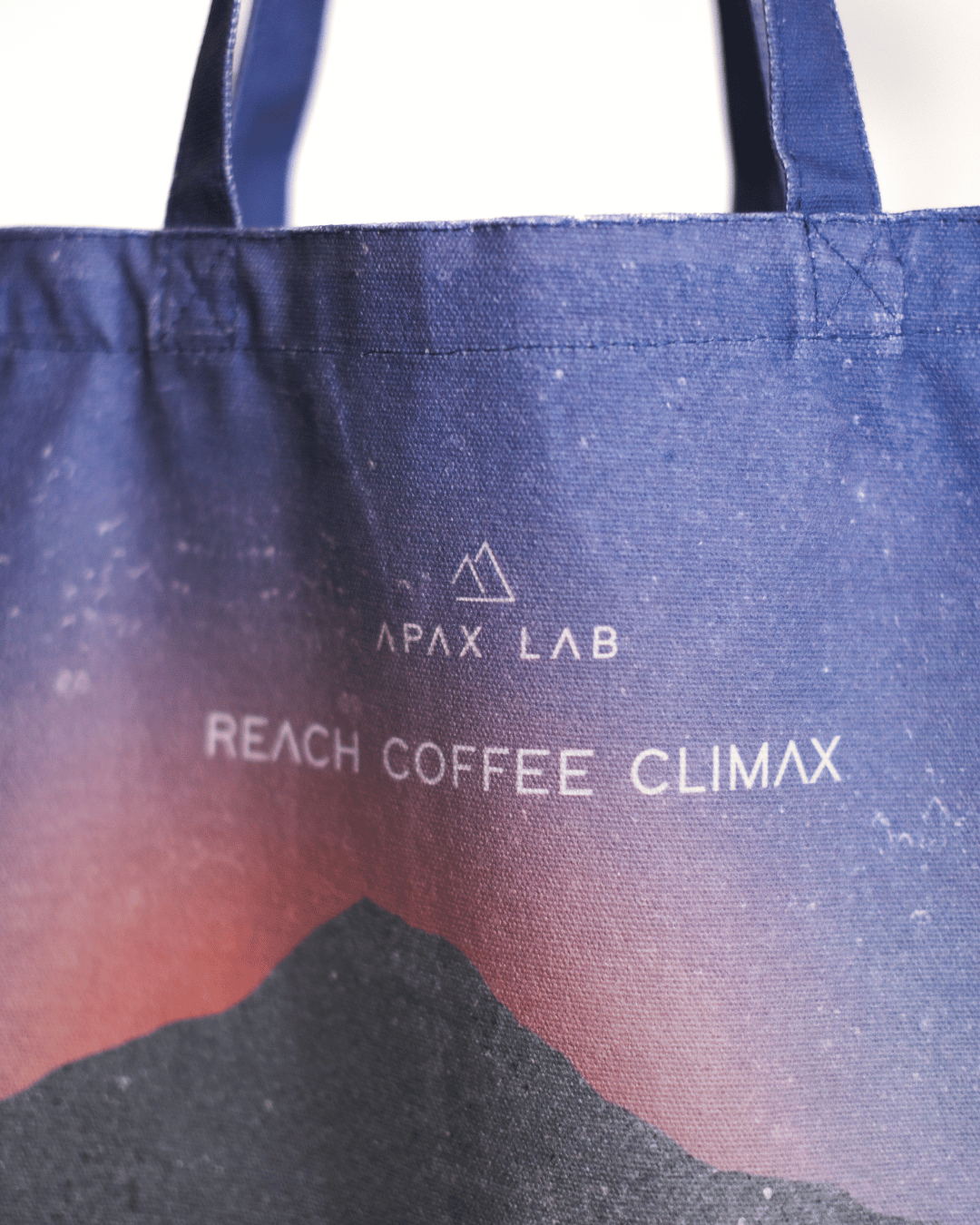 A close-up of a blue canvas tote bag with white printed text reading 'APAX LAB' and 'REACH COFFEE CLIMAX,' plus a mountain graphic near the bottom. The bag fills most of the frame against a light neutral background.