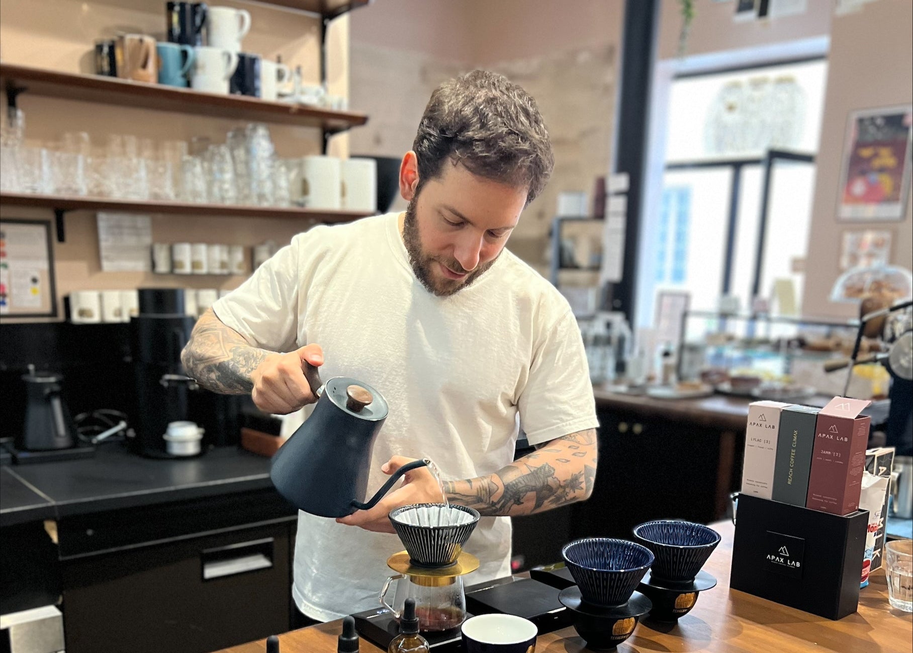 Barista preparing coffee in a coffee shop with shelves and equipment in the background.